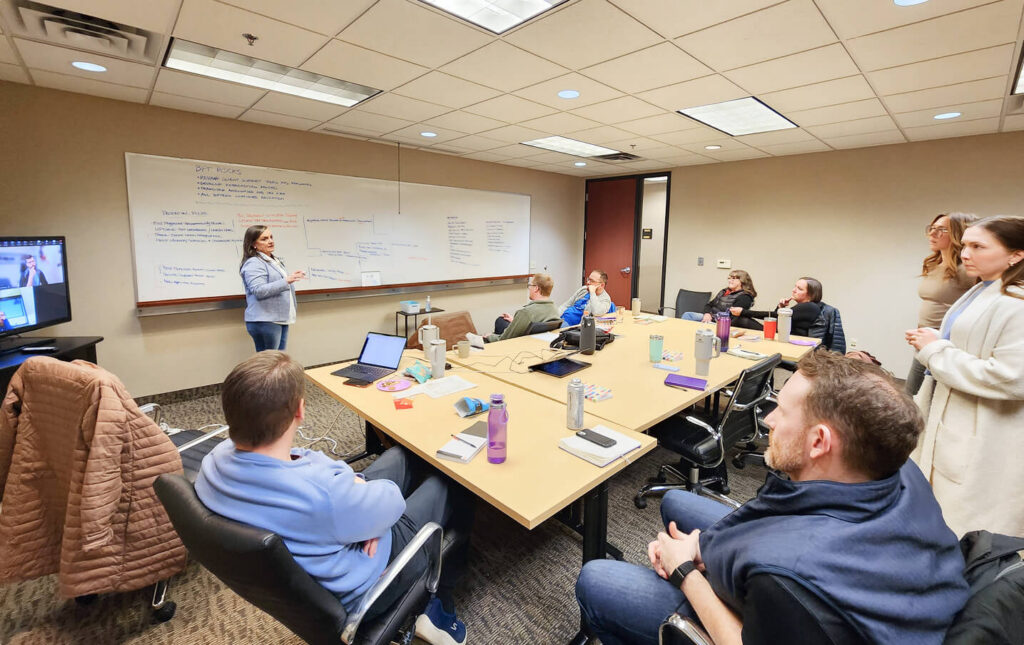A woman stands and presents to a group of people seated around a conference table in an office. Notes are written on a large whiteboard, and laptops, water bottles, and notebooks are on the table.