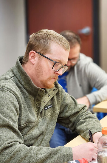 A man with glasses and a green fleece jacket writes on paper at a desk in a classroom. Another person in a gray hoodie is seated behind him, also writing. The focus is on the man in front.