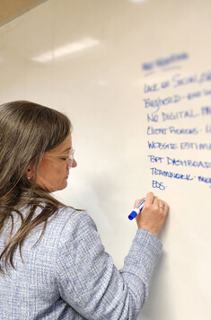 A woman with long brown hair and glasses writes with a blue marker on a whiteboard covered in blurred handwritten text. She is wearing a light gray blazer.