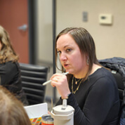 A woman with straight brown hair, wearing a black top, sits at a table holding a white pen near her mouth, looking toward the camera. A drink cup and papers are on the table in front of her.